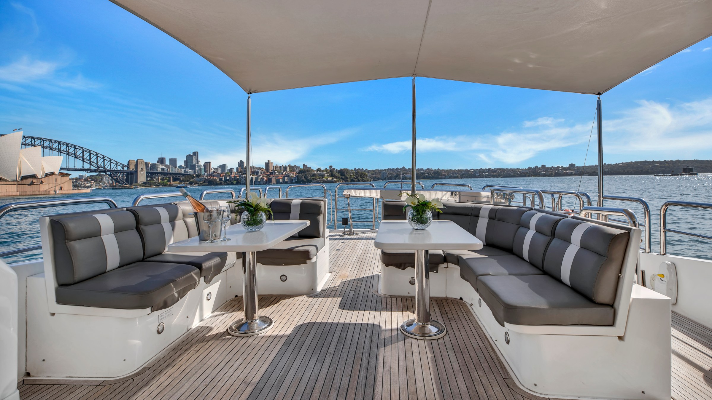 Luxury boat deck with seating, tables, and city skyline with bridge and opera house in background.
