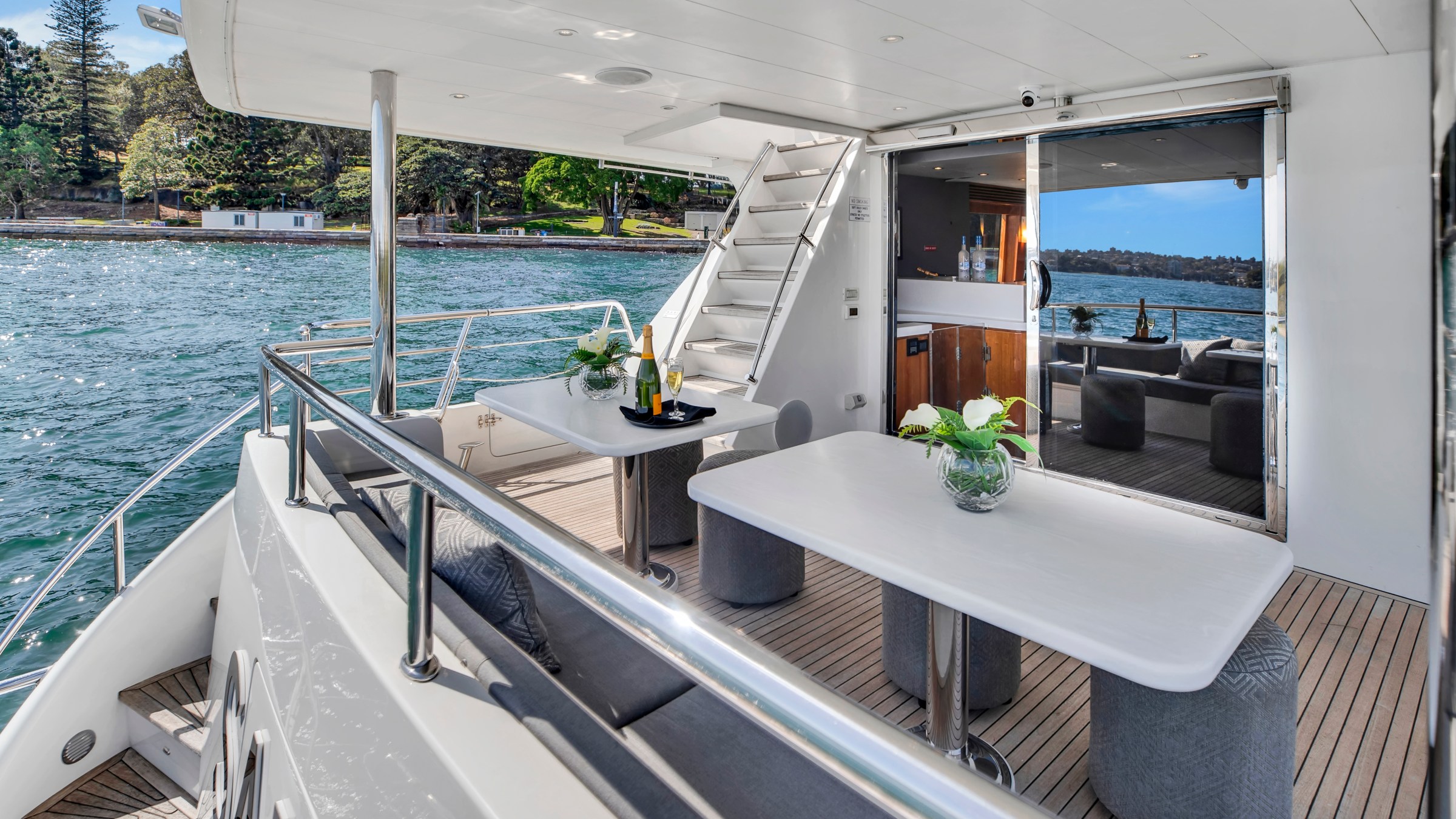 Luxury yacht deck with tables, flowers, and water view. Sliding doors lead to interior.
