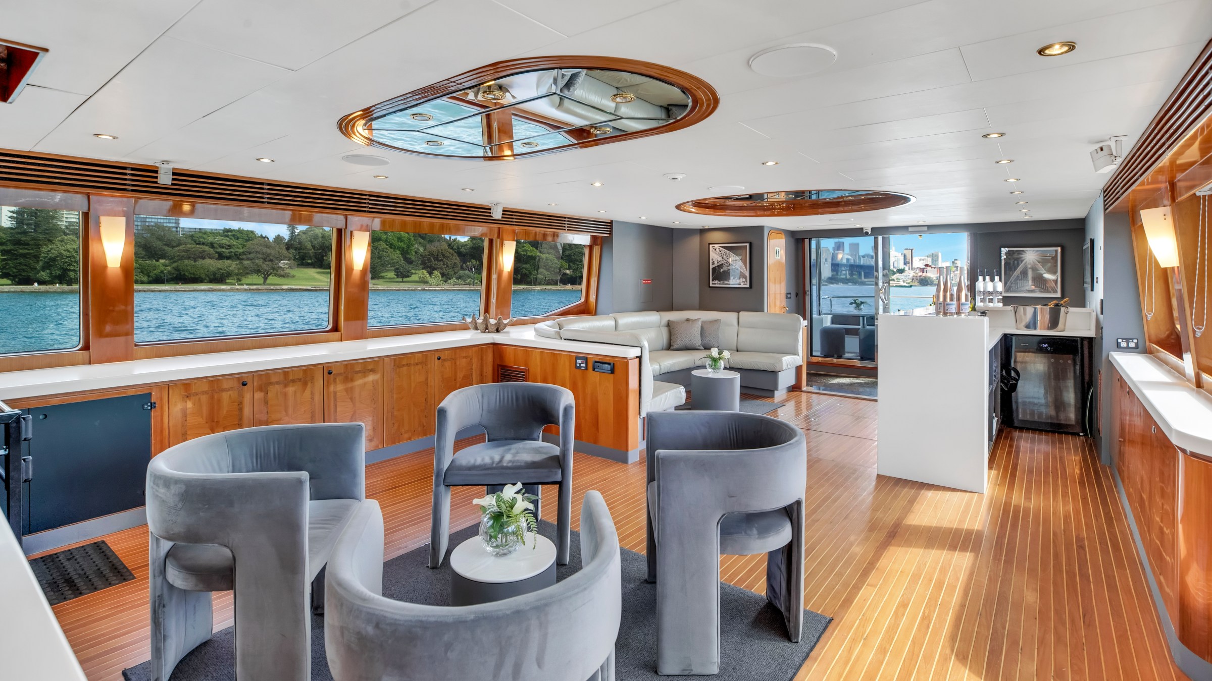 Luxury yacht interior with gray chairs, wooden flooring, large windows, and a scenic water view.