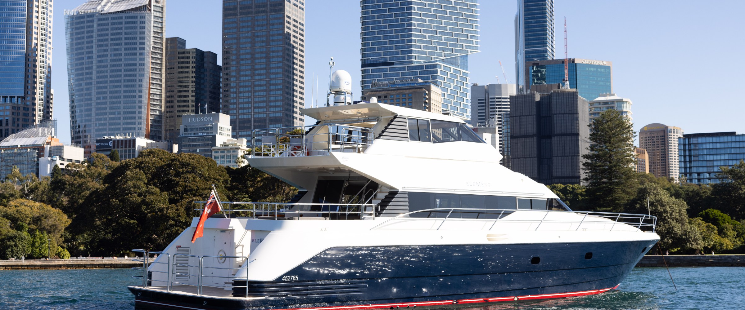 Luxury yacht on water with city skyline and trees in the background.