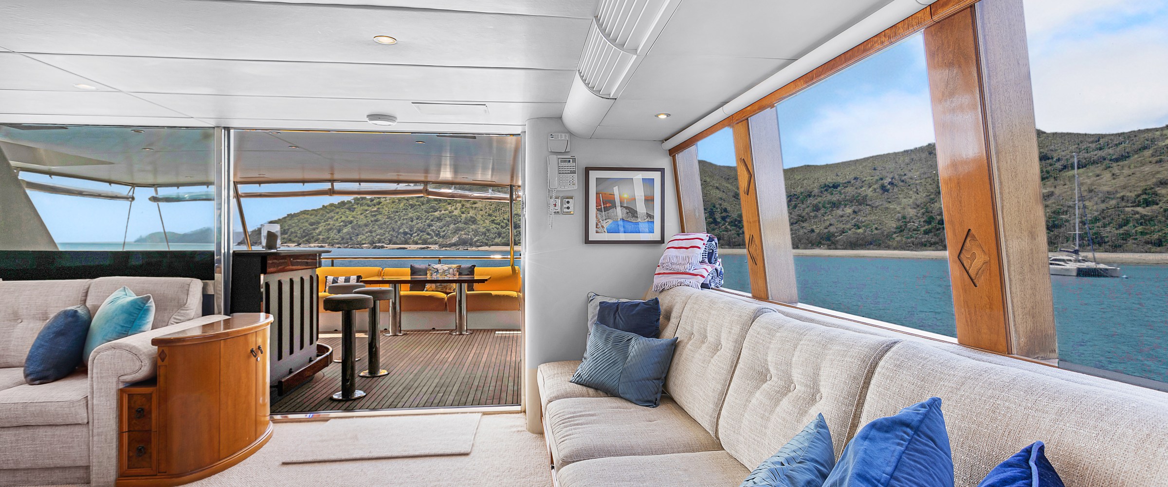 Spacious yacht interior with large windows, beige couches, and blue pillows overlooking a scenic ocean view.