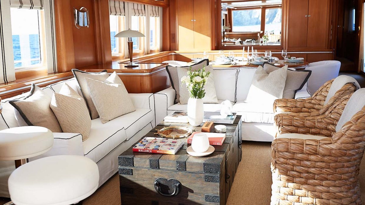 Luxurious yacht interior with white sofas, wicker chairs, books on coffee table, and dining area.