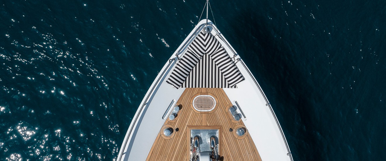 Aerial view of a yacht's bow with striped cushions on wood deck, surrounded by blue ocean water.