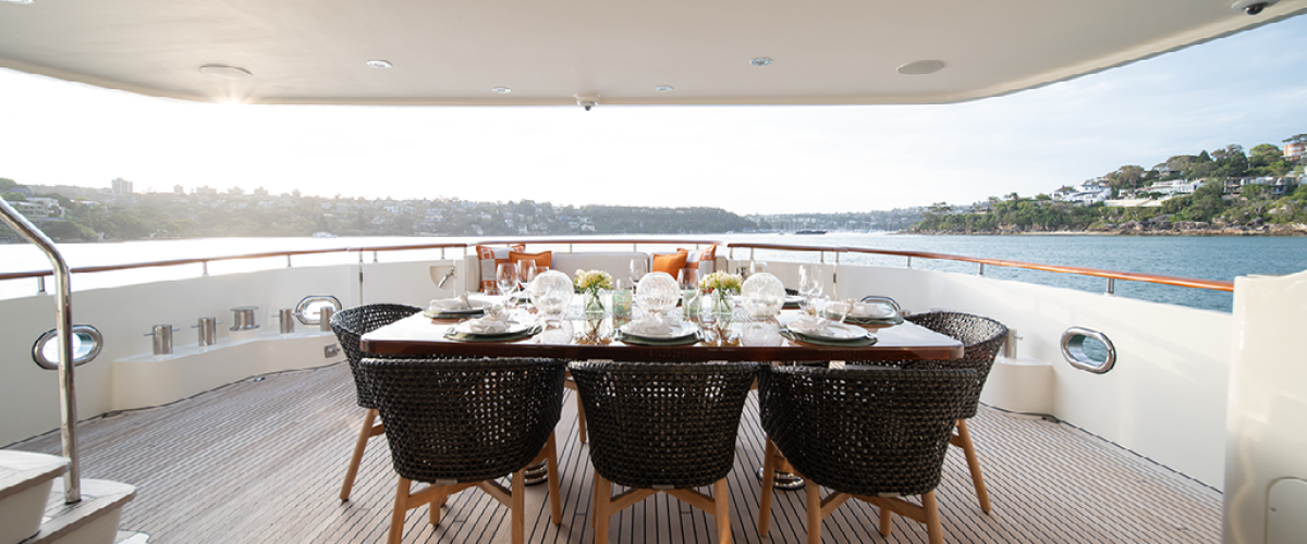 Outdoor dining setup on yacht deck with sea view and distant hills.