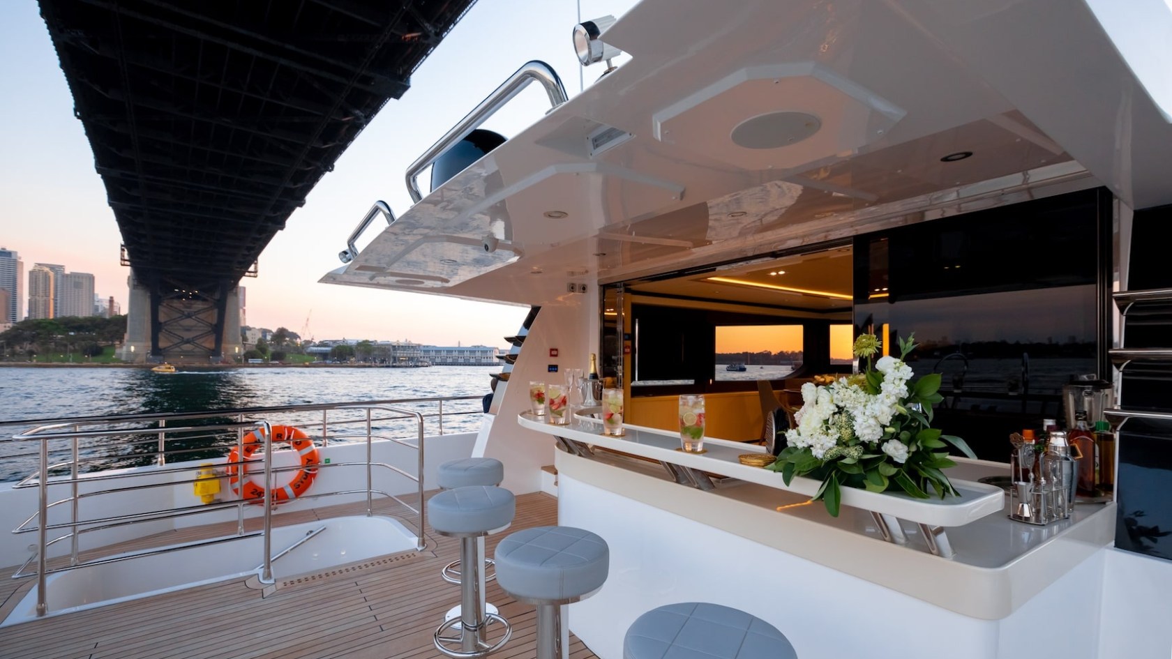Luxury yacht deck with bar, stools, and floral arrangement under a large bridge at sunset.