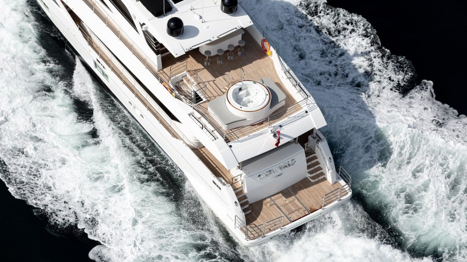 Aerial view of a white luxury yacht cruising through waves with a wooden deck and hot tub on the stern.