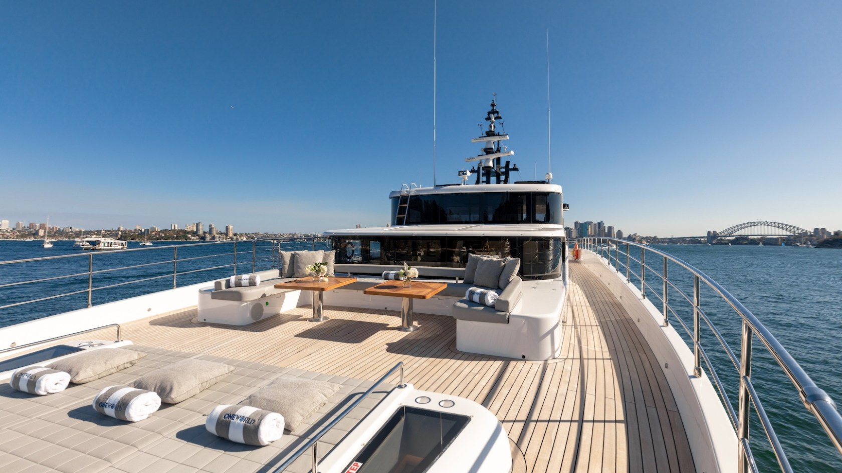Luxury yacht deck with seating and tables, on a sunny day with city skyline and bridge in background.