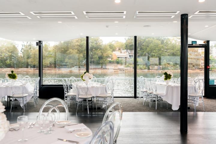 Elegant dining area with round tables and clear chairs, overlooking a river view through large windows.