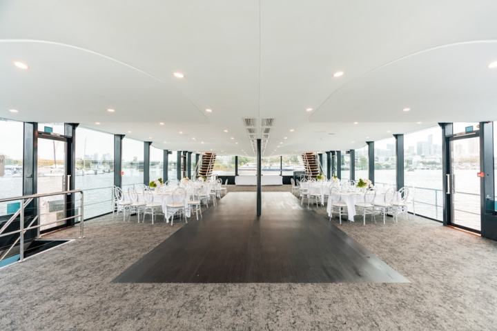 Spacious interior of a boat with dining tables, chairs and large windows overlooking water.