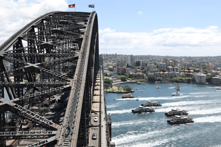 Sydney Harbour Bridge with flags, cars, and boats in the water below on a sunny day.