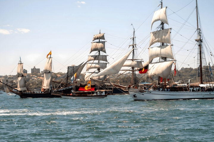 Three tall ships with sails unfurled on a sunny day in a harbor.