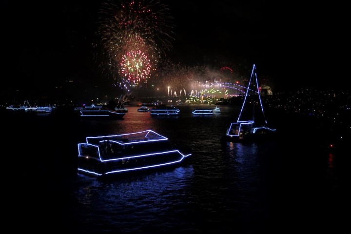 Boats with neon lights in a harbor with colorful fireworks in the sky.