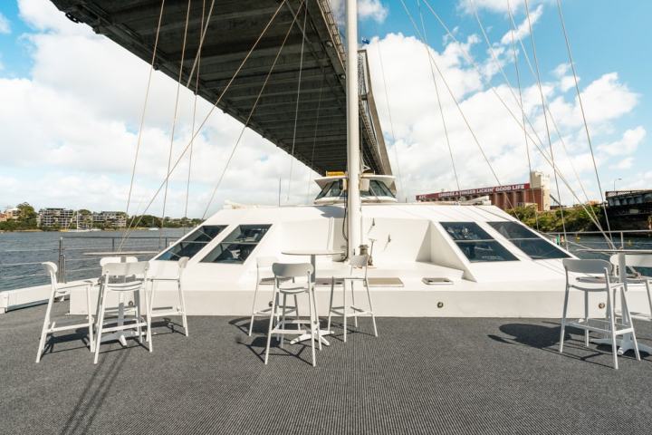 Deck of a boat under a bridge with tables and chairs, city in background.