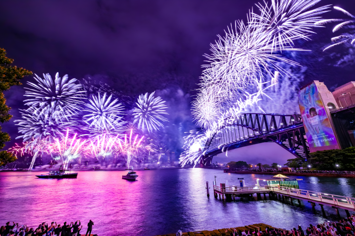 Spectacular fireworks over a bridge at night with reflections on the water and a crowd watching.
