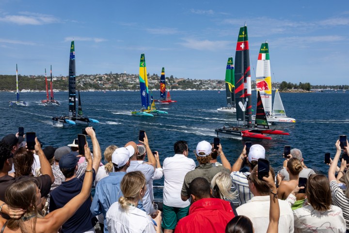 Crowd watches sailboat race with colorful sails in a bay on a sunny day.