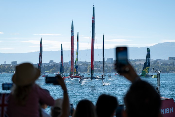 People photograph sailboats competing in a race on a sunny day with mountains in the background.