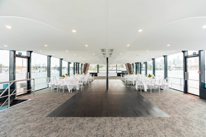 Spacious event hall on a boat with white chairs and tables, large windows, and a view of the water.