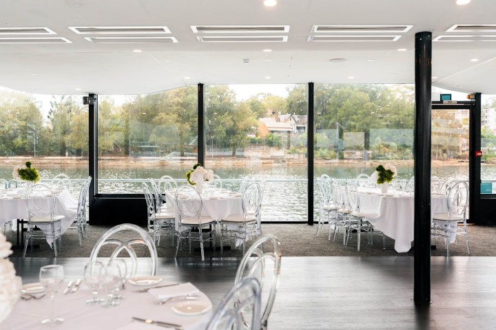 Elegant dining area with round tables, glass chairs, white linens, and large windows facing a riverside view.