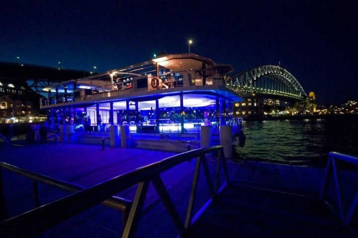 Night view of a lit-up dockside building near a bridge over a body of water.